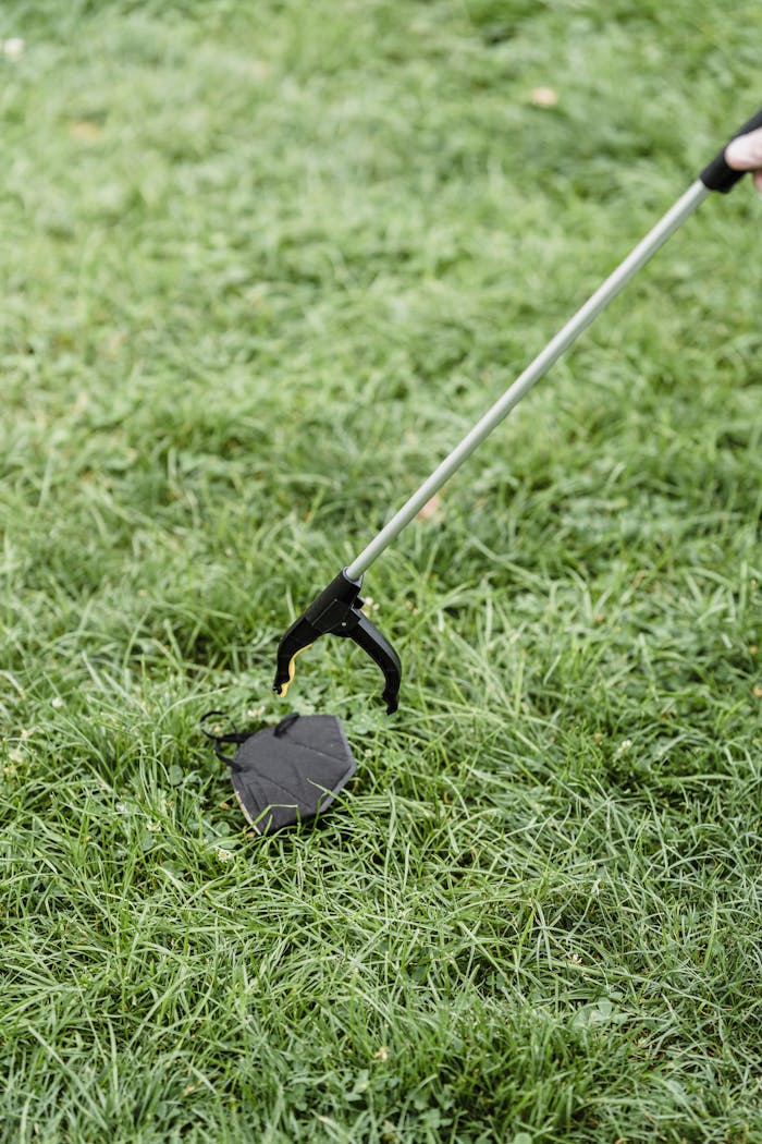 A litter picker tool collects a discarded face mask from a grassy lawn, emphasizing environmental concerns.
